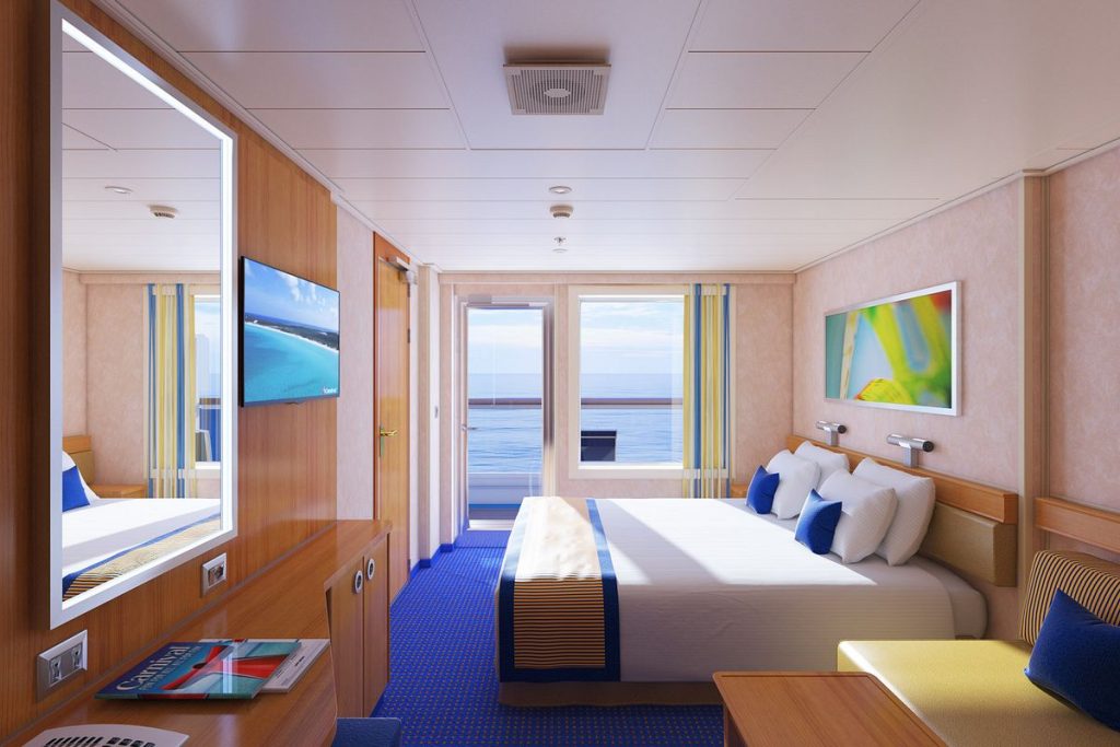 Carnival Radiance balcony room with a queen-size bed, modern decor featuring blue and gold tones, and a sliding door leading to a private balcony with ocean views