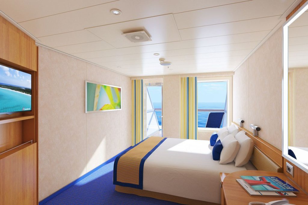 Aft-View Extended Balcony stateroom on Carnival Radiance, featuring a queen bed, contemporary decor with blue carpet, and a sliding door to a private balcony with stunning ocean views.