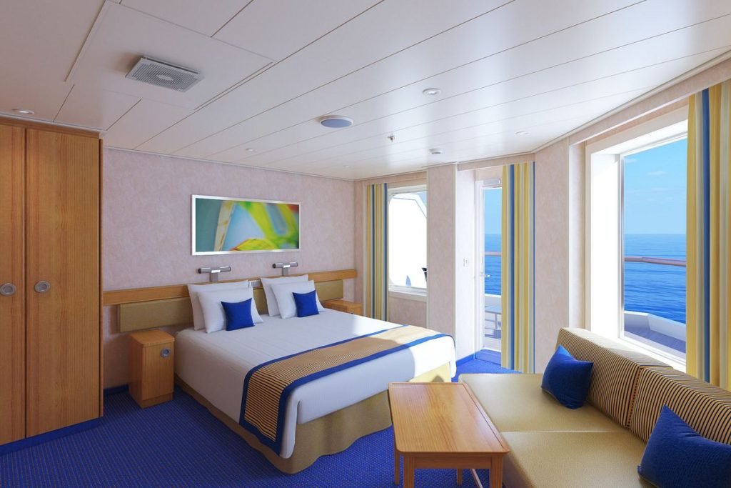 This image showcases a bright and cozy balcony cabin on a Carnival cruise ship. The room features a comfortable bed with white linens and blue accent pillows, a small seating area with a sofa and table, and large windows leading to a private balcony with ocean views. The design is cheerful and functional, perfect for relaxing while enjoying the sea breeze.