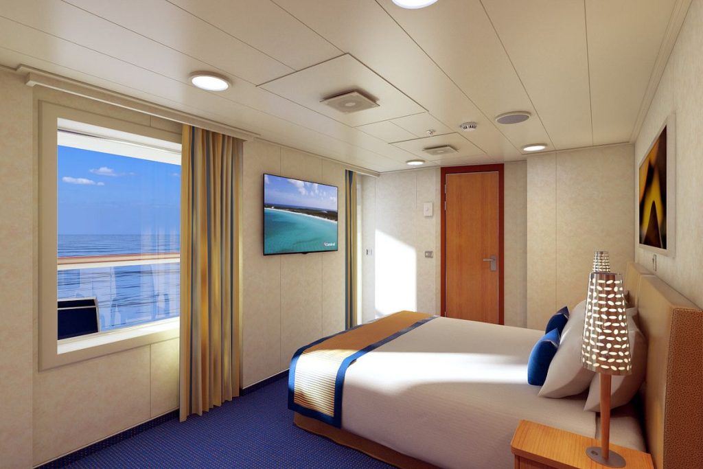 Interior view of a Carnival cruise ship premium balcony room with a large bed, a mounted flat-screen TV, and a window showing a serene ocean view.