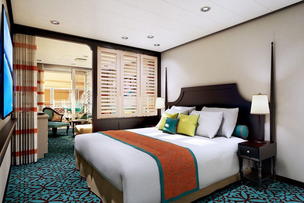 Elegant Havana Cabana Suite on Carnival Vista with a large bed adorned with white linens and a vibrant orange and teal runner. The room features a tropical patterned carpet, wood-finished bedside tables, and decorative pillows that add a pop of color. Slatted doors open to a private cabana area, inviting natural light and offering a glimpse of the outdoor seating and hammock.