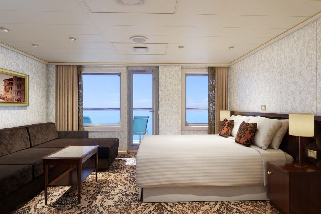 A tastefully decorated Ocean Suite on the Carnival Venezia, featuring a large bed with striped bedding and accent pillows, a plush brown sofa, ornate carpeting, and a framed picture above a side table, all with a view of the sea through two windows.
