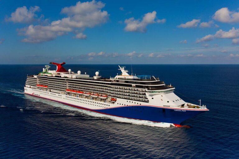 The Carnival Pride cruise ship journeys near Grand Cayman, its distinctive red and blue funnel a vibrant contrast against the bright blue sky. The ocean's deep blues surround the vessel, emphasizing its elegant white structure as it cruises the serene Caribbean Sea.