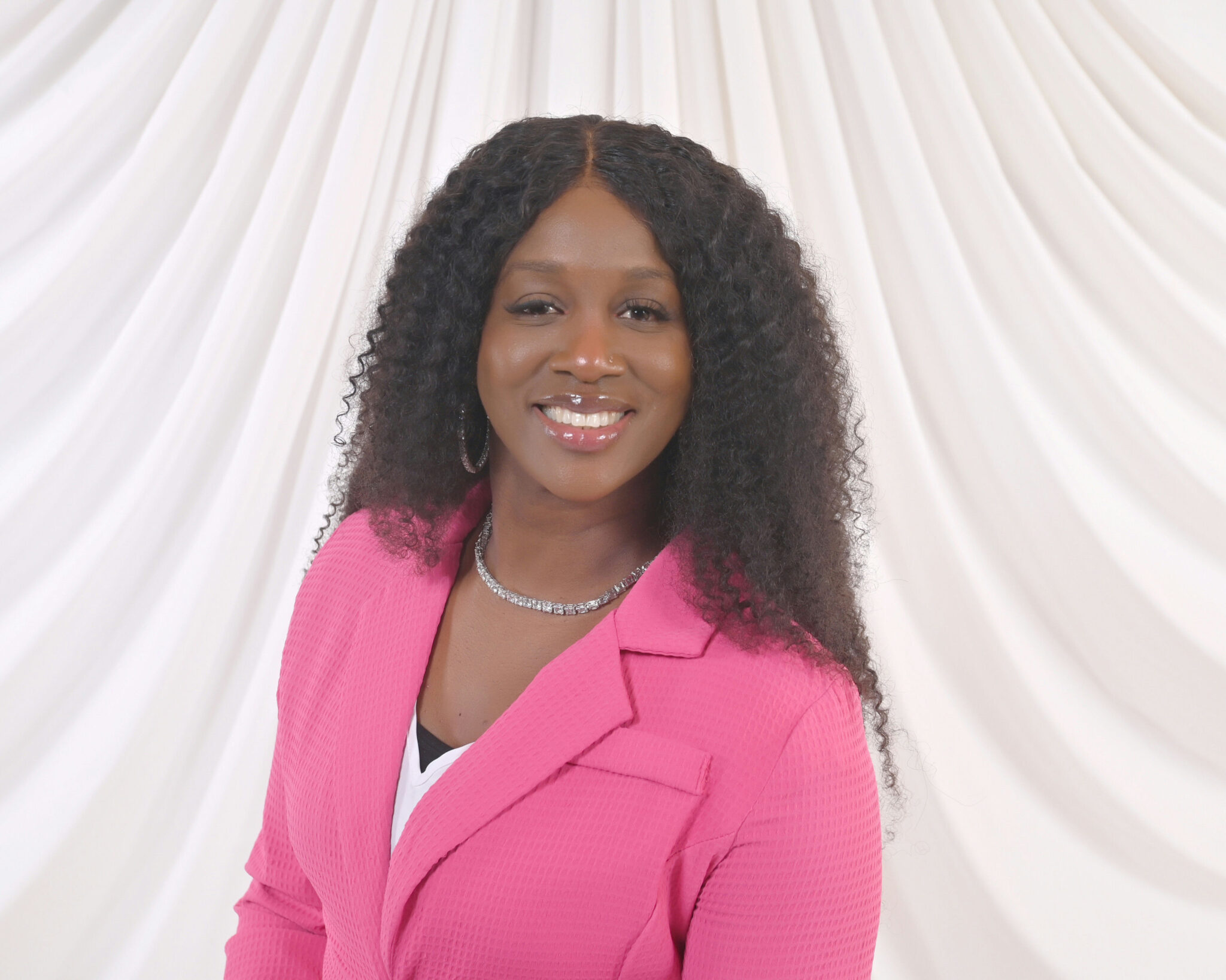 Cryssi St. Cyr, a Carnival cruise director, smiles confidently while posing in front of a white draped backdrop. She is wearing a bright pink blazer, a white top, and a sparkly necklace, presenting a professional and approachable demeanor.