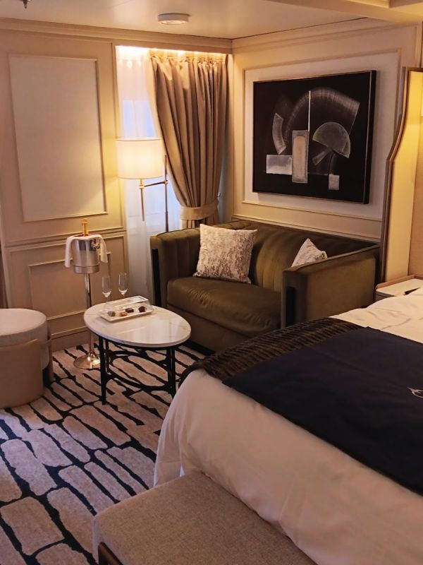 This image depicts a luxurious cabin on the Oceania Mariner cruise ship. The room features an elegant sofa with plush pillows, a small round coffee table with champagne glasses, and a tray of refreshments. A stylish, modern piece of artwork is mounted on the wall, and a nearby floor lamp provides warm lighting. The room also has a cozy bed with a dark throw, creating an inviting atmosphere. The soft, neutral color palette and sophisticated decor offer a relaxing, upscale ambiance.