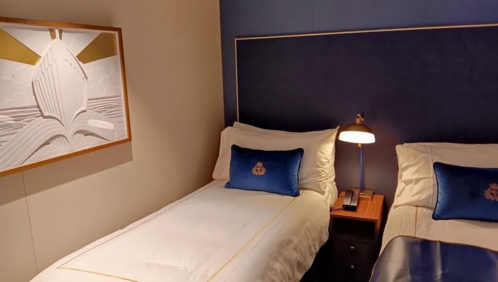 The image shows a cozy cabin on Cunard's Queen Anne cruise ship. The room features two neatly made single beds with white linens and blue accent pillows, each embroidered with a gold crest. A bedside table between the beds holds a classic reading lamp, adding a warm glow to the space. On the wall, a framed piece of modern artwork depicts a ship with stylized waves and sun rays in a minimalist design, enhancing the nautical theme of the room. The atmosphere is both elegant and comfortable, with a simple yet refined decor.