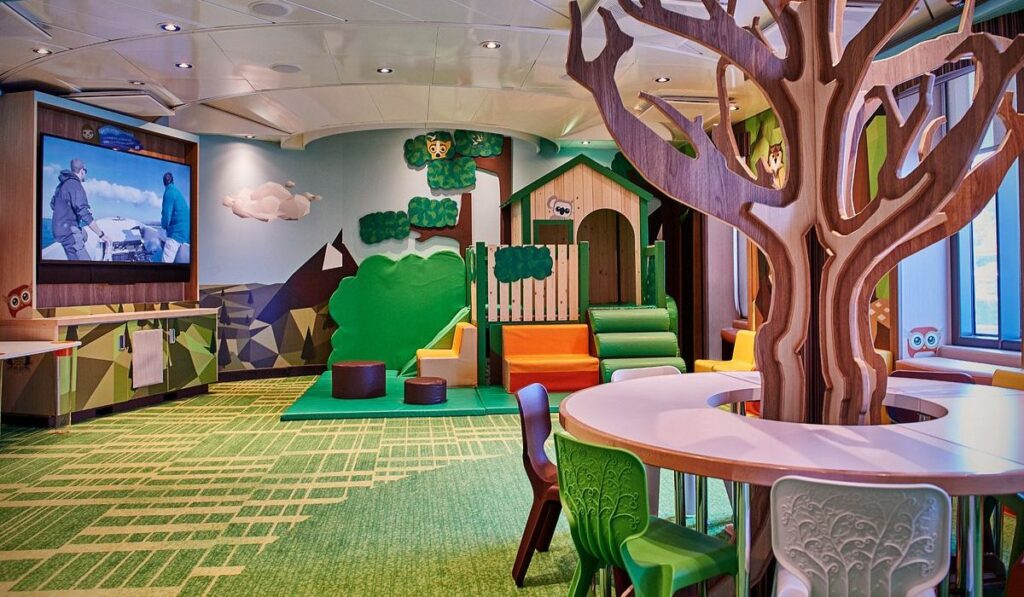 The image shows Camp Discovery, a children's play area on a Princess Cruise ship, designed to engage and entertain young cruisers. The vibrant room boasts a whimsical, nature-inspired decor with a large tree structure, playful animal motifs, and a colorful playhouse. Brightly colored seating and an interactive floor pattern create an inviting environment for children to play and learn. The large screen on the wall and the windows offering a view of the sea suggest an integration of technology and scenic enjoyment, enhancing the interactive experience for kids.