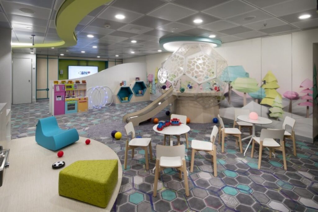 This image showcases the "Camp at Sea" kids' play area aboard the Celebrity Edge cruise ship. The space is creatively designed with colorful, interactive elements, including a small slide, a modern dome-shaped climbing structure, and hexagonal cubby spaces. The room also features child-sized tables and chairs, playful wall art, and a variety of toys, providing a fun and engaging environment for younger guests.