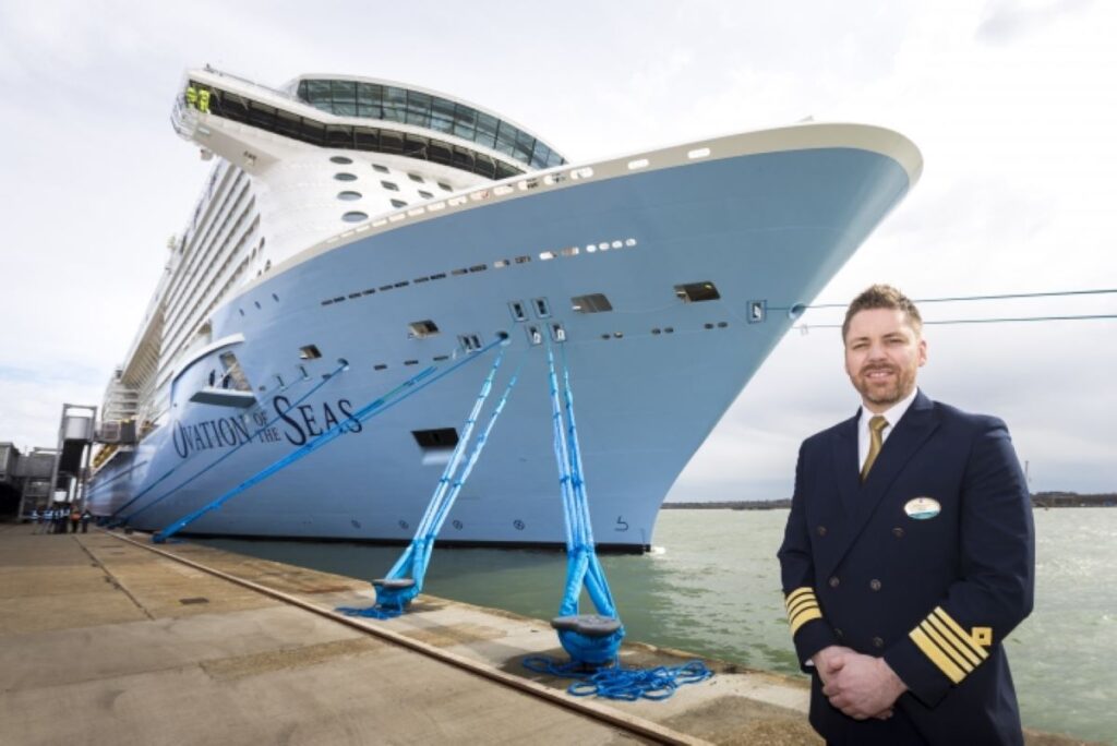 Captain Henrik Loy stands proudly in front of Royal Caribbean's Ovation of the Seas, with the massive cruise ship's hull towering in the background, moored with thick blue ropes, illustrating the grand scale of the vessel and the esteem of its captain.
