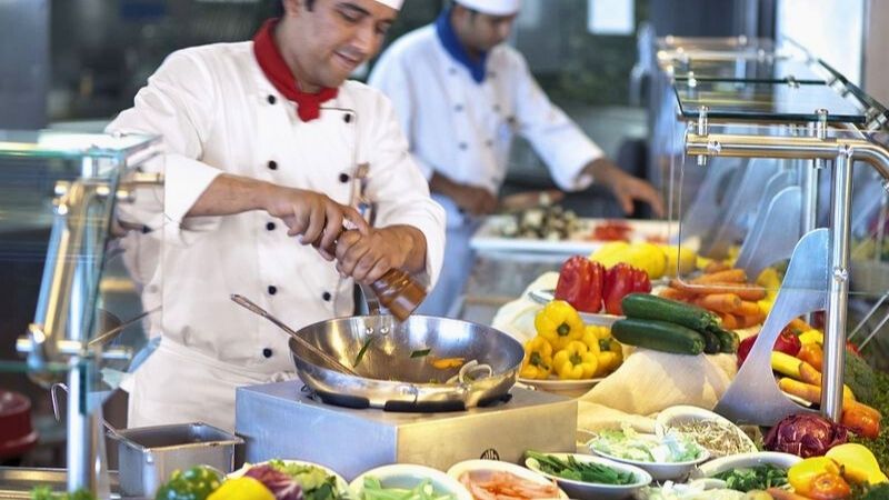 A chef in a white uniform and red neckerchief seasons a stir-fry dish at a live cooking station, surrounded by a colorful array of fresh vegetables including bell peppers, carrots, zucchini, and greens. Another chef in the background prepares food at a separate station, highlighting the vibrant, bustling atmosphere of a buffet-style kitchen.
