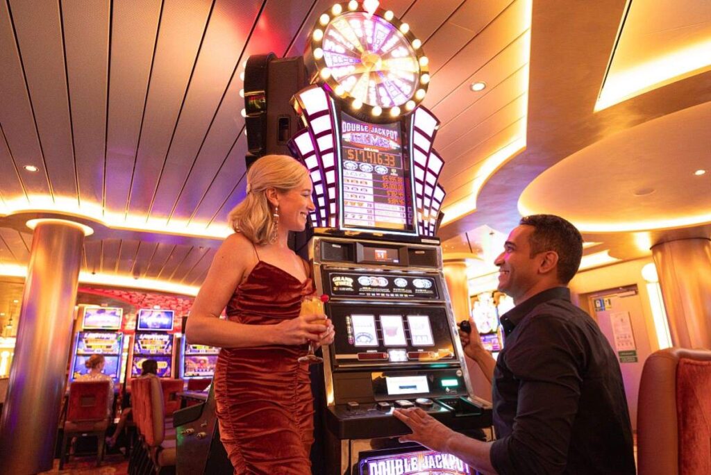 A woman in an elegant red velvet dress smiles while playing a slot machine, with a man next to her looking on joyfully, in the vibrant and lively ambiance of the Carnival casino, surrounded by colorful gaming machines and soft overhead lighting.
