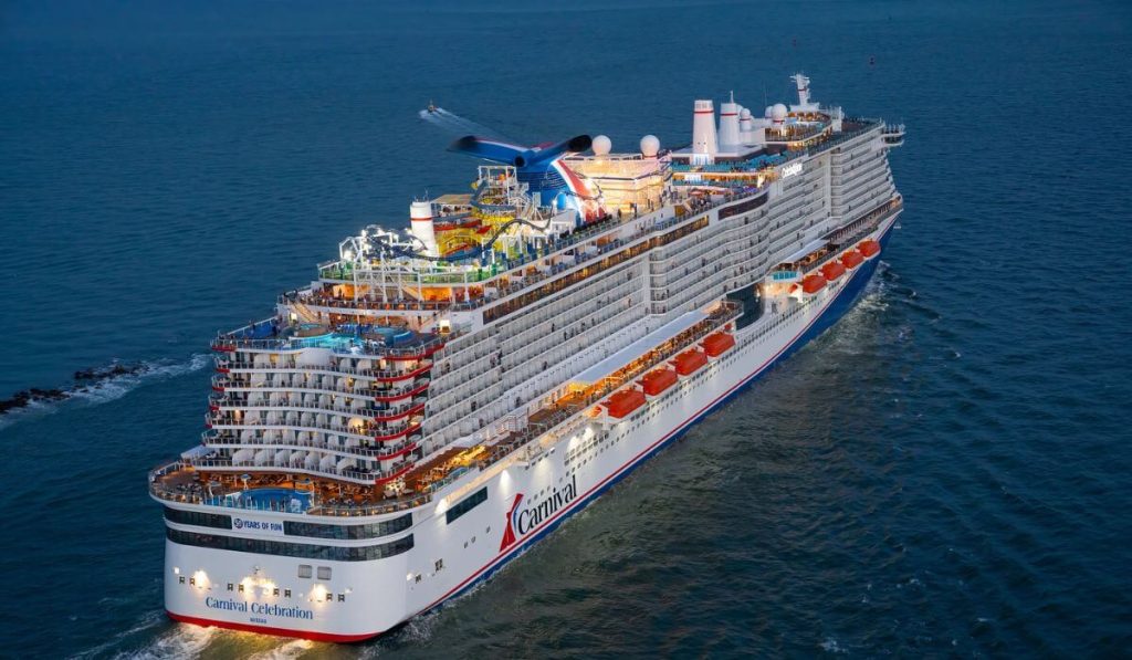 The Carnival Celebration cruise ship is seen from above, showcasing its multiple decks with colorful water slides and leisure amenities, making its way through the blue ocean.