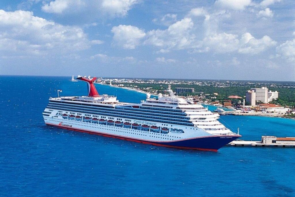 Carnival Cruise Line's Carnival Conquest is pictured docked in a bustling port with the clear turquoise sea stretching before it. The ship's distinctive red and blue funnel stands out against the coastal town backdrop, showcasing the vibrant colors and leisurely atmosphere of a tropical destination.
