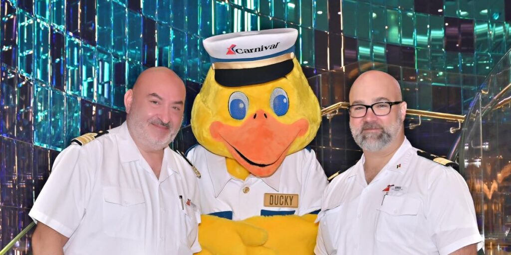 This image features two individuals in white uniforms with epaulets, posing alongside a large yellow duck mascot dressed in a sailor's hat and uniform with the Carnival logo and a name tag that reads "Ducky." The background is a reflective, colorful setting that suggests it might be aboard a cruise ship or a festive environment.