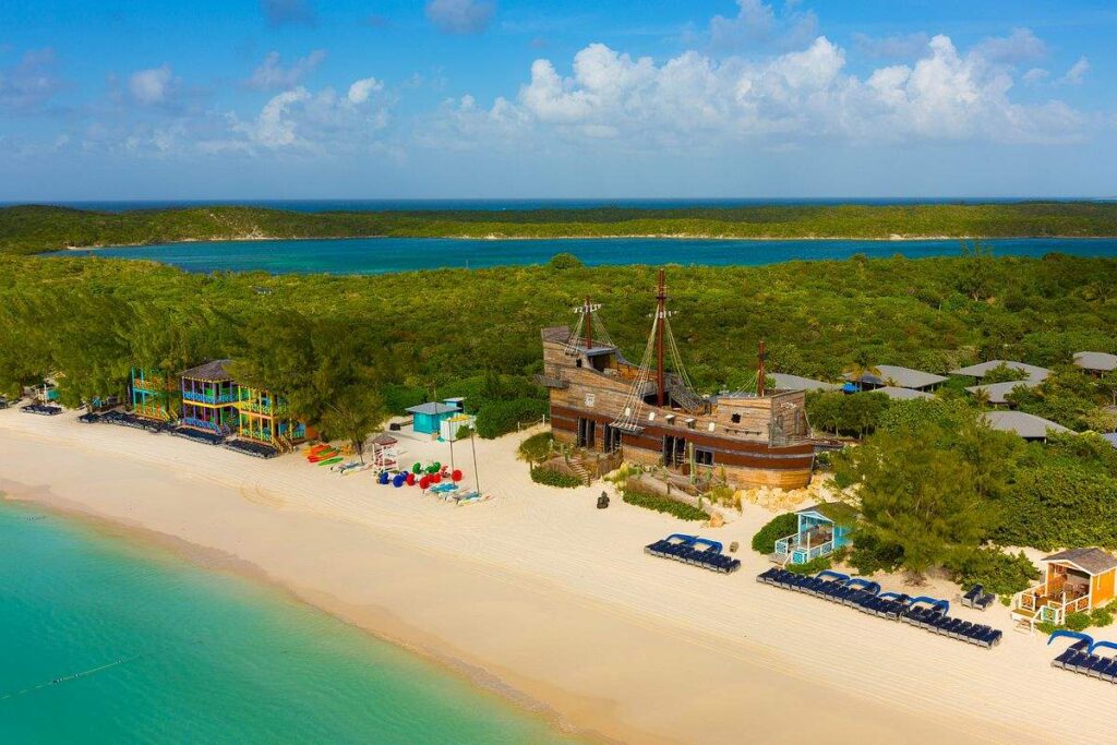 Aerial view of Half Moon Cay, Carnival's private island, showcasing its white sandy beach with sun loungers, vibrant blue waters, and a unique pirate ship structure amid lush greenery.