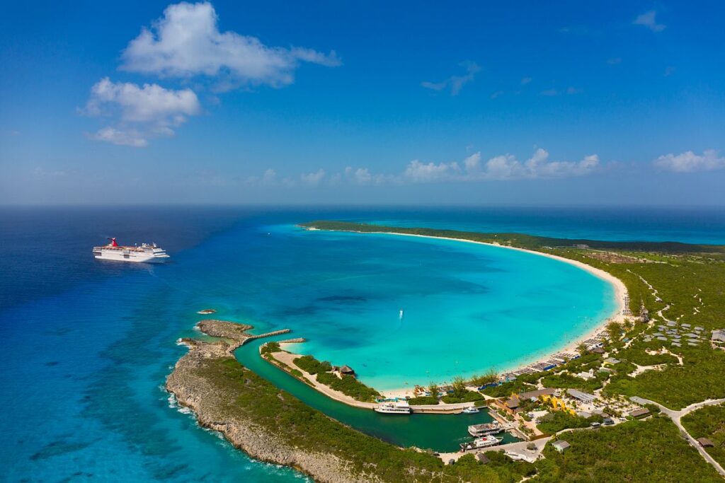 Carnival Elation cruise ship anchored off the coast of Half Moon Cay, featuring the ship's iconic red funnel against the stunning backdrop of the island's crescent-shaped white sand beach and turquoise waters.