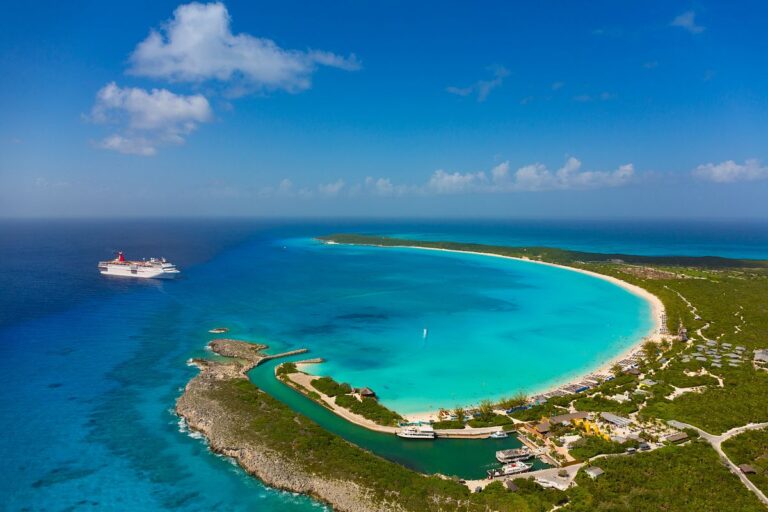 Carnival Elation cruise ship anchored off the coast of Half Moon Cay, featuring the ship's iconic red funnel against the stunning backdrop of the island's crescent-shaped white sand beach and turquoise waters.