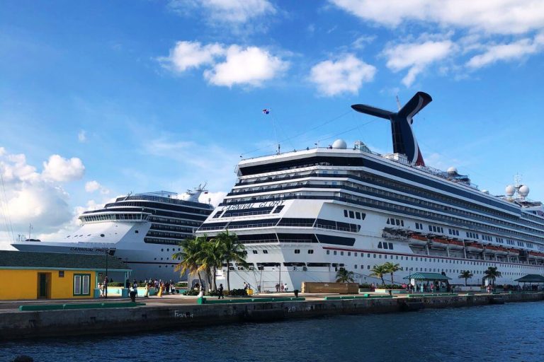 Carnival Glory and Carnival Pride cruise ships docked side by side under a clear blue sky, with passengers strolling on the quay.