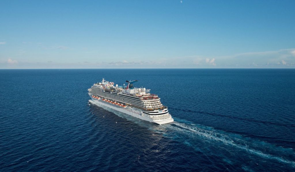 The Carnival Horizon cruise ship sails through calm, sapphire-blue waters under a clear sky, with the ship's multiple decks and signature red funnel visible from an aerial perspective.