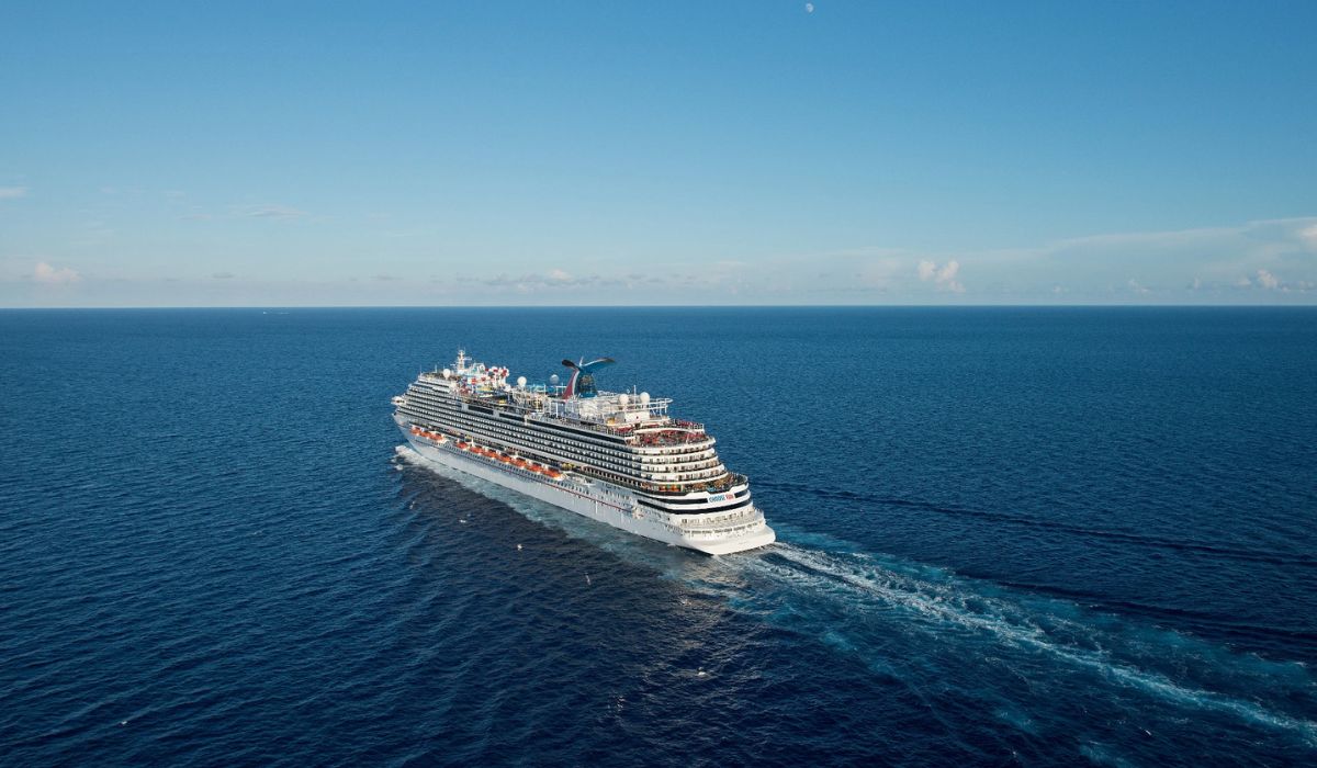 A majestic cruise ship, possibly the Carnival Horizon, glides across the serene deep blue waters of the ocean, its multiple decks and amenities highlighted under the vast, cloud-streaked sky. The vessel carves a frothy trail in the sea, showcasing the grandeur of maritime travel.