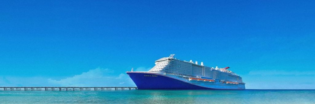 The Carnival Jubilee cruise ship is pictured docked near a long pier, with the turquoise sea in the foreground and a bright blue sky above.