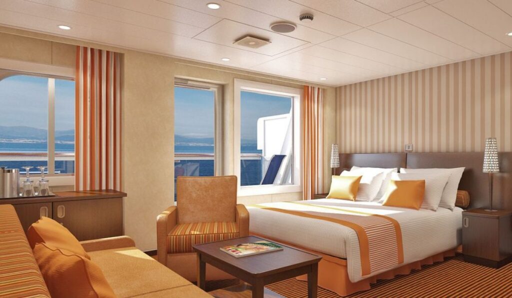 The Junior Suite on Carnival Liberty, tastefully decorated in warm earth tones with a large bed accented by orange and yellow throw pillows. A comfortable sitting area and expansive windows offer a serene view of the sea, making the space ideal for relaxation and enjoyment of the cruise experience.