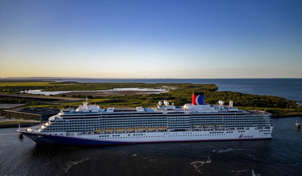 The image showcases the Carnival Luminosa cruise ship elegantly docked in a tranquil waterway, with lush coastal greenery in the background and a serene sky above as daylight fades.
