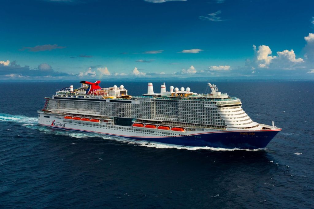 The Carnival Mardi Gras cruise ship cuts through the deep blue sea, its towering red and white funnel standing out against a backdrop of scattered clouds in a vibrant blue sky.