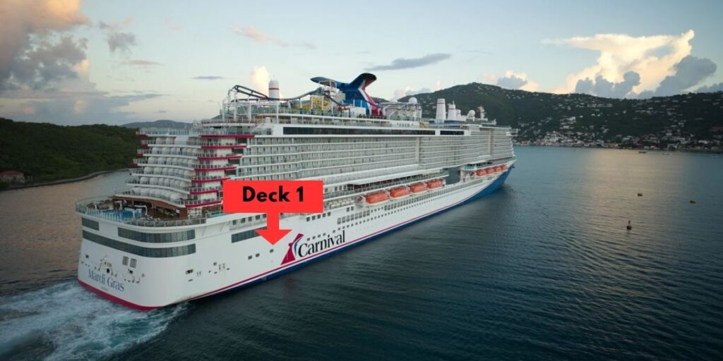 This image shows the Carnival Mardi Gras cruise ship sailing in calm waters with lush, green hills and a coastal town visible in the background. The cruise ship is large and modern, with an exterior that prominently displays the Carnival logo. A red graphic is added to the image, indicating the location of "Deck 1" with a bold arrow pointing to the lower part of the ship. The sky is partly cloudy, creating a serene setting.
