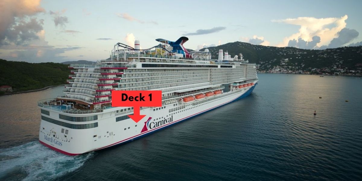 This image shows the Carnival Mardi Gras cruise ship sailing in calm waters with lush, green hills and a coastal town visible in the background. The cruise ship is large and modern, with an exterior that prominently displays the Carnival logo. A red graphic is added to the image, indicating the location of "Deck 1" with a bold arrow pointing to the lower part of the ship. The sky is partly cloudy, creating a serene setting.