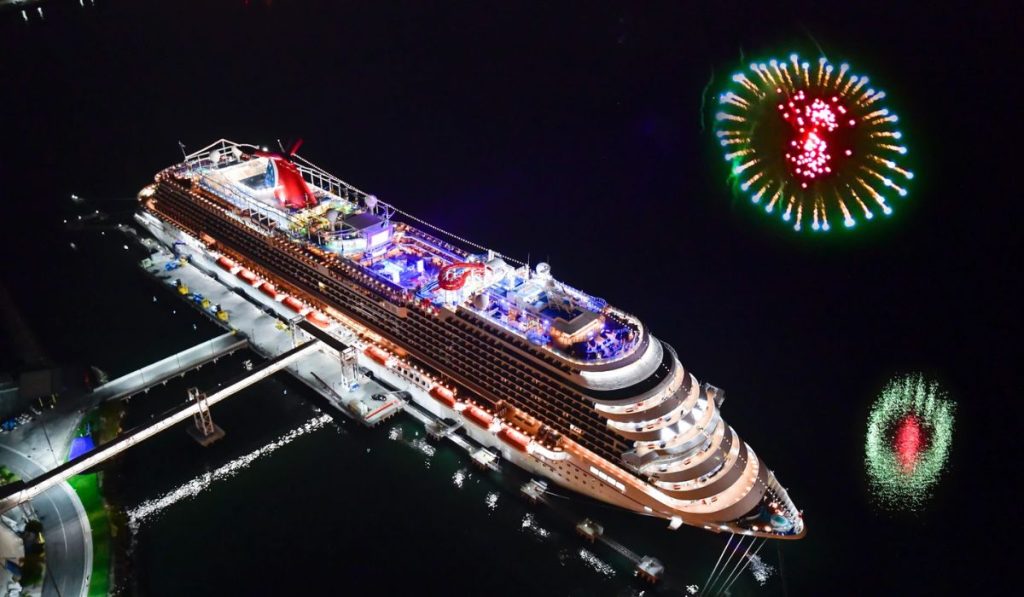 Nighttime aerial view of the Carnival Panorama cruise ship docked at port, brilliantly illuminated with festive lights, with a backdrop of vibrant fireworks decorating the dark sky.