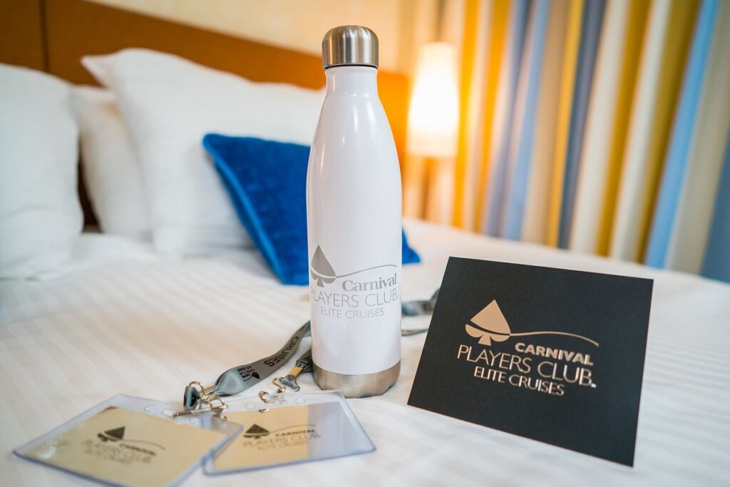 A welcoming setup in a Carnival cruise cabin with 'Carnival Players Club Elite Cruises' branded amenities, including a white insulated bottle, key cards on a lanyard, and a matching information card, all arranged on a crisp white bedspread with a striped headboard and cozy pillows in the background.