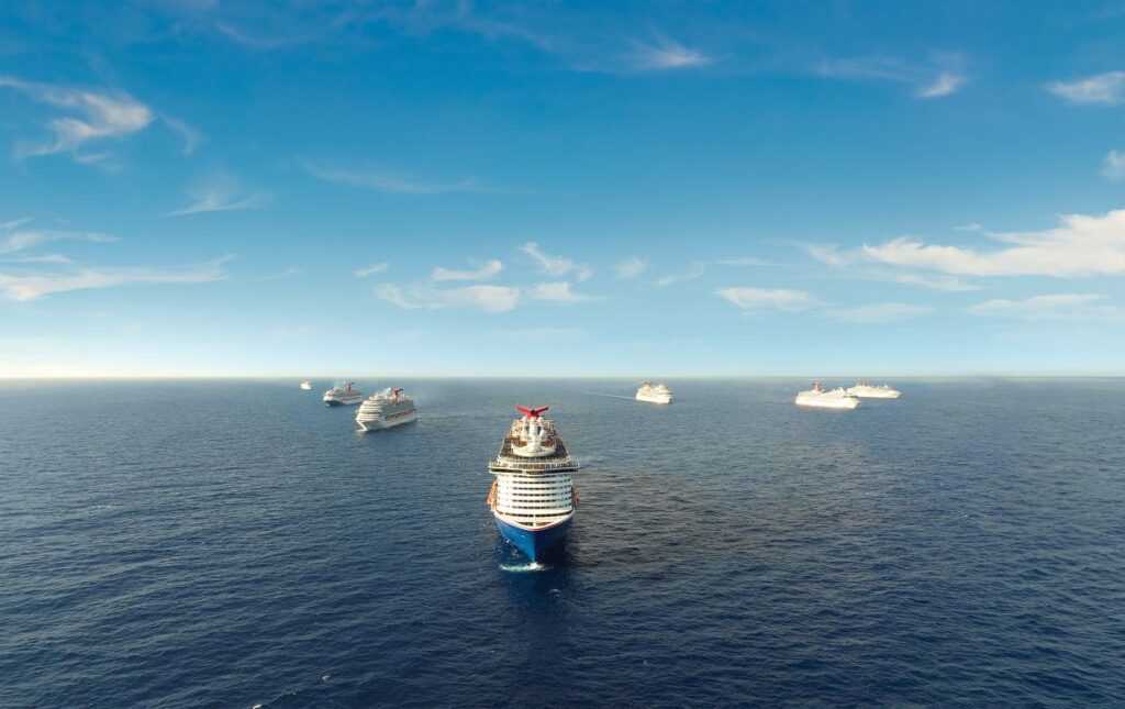 A fleet of Carnival cruise ships gathers in the tranquil blue waters of the Bahamas under a vast sky with scattered clouds.