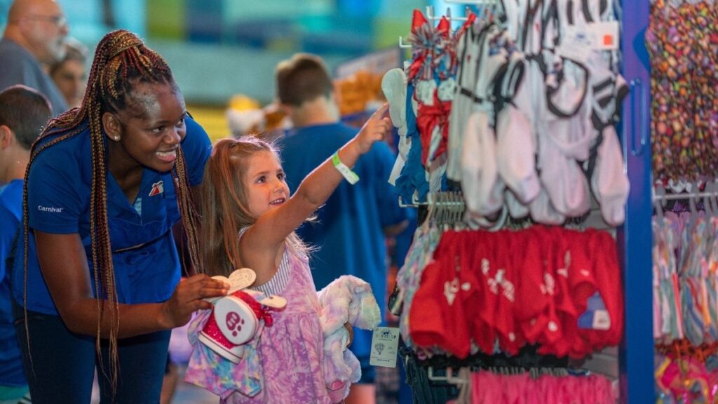 A photo in a Carnival cruise gift shop, of a young girl choosing something to buy and a member of crew helping her.