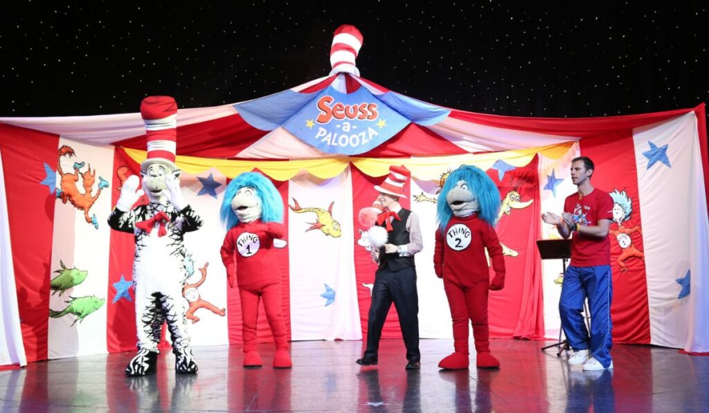 Suess-a-Palooza Storytime and Parade on a Carnival Cruise, with characters from Dr. Seuss's books like the Cat in the Hat and Things 1 and 2 engaging with an audience against a whimsical stage backdrop.