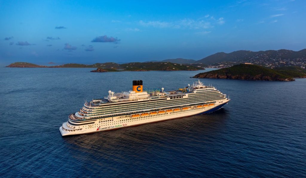 The Carnival Venezia cruise ship is captured in the golden light of dusk, cruising near hilly coastlines with scattered residential areas.