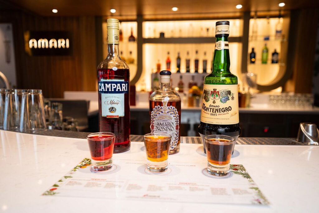 The image showcases an elegant Amari bar onboard the Carnival Venezia, featuring three prominent bottles of Italian liqueurs — Campari, Amaro Nonino, and Amaro Montenegro — in sharp focus on a marble countertop. In the foreground, three glasses of the amari are neatly lined up, suggesting a tasting is about to take place. The warm ambiance of the bar provides a sophisticated backdrop for these classic Italian spirits.