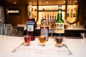 The image showcases an elegant Amari bar onboard the Carnival Venezia, featuring three prominent bottles of Italian liqueurs — Campari, Amaro Nonino, and Amaro Montenegro — in sharp focus on a marble countertop. In the foreground, three glasses of the amari are neatly lined up, suggesting a tasting is about to take place. The warm ambiance of the bar provides a sophisticated backdrop for these classic Italian spirits.