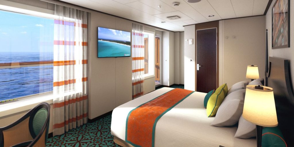 An inviting stateroom onboard the Carnival Horizon, featuring a large bed with a vibrant orange throw, a comfortable seating area, and a flat-screen TV displaying a beach scene. The room is bathed in natural light from the ocean-view window, creating a serene and cozy atmosphere for guests.