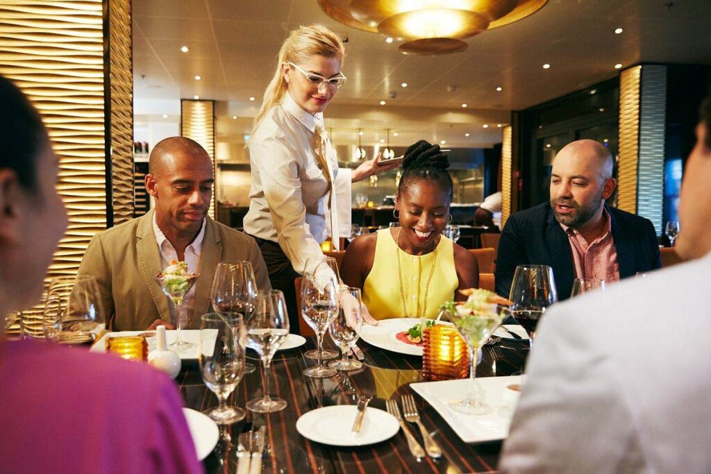 The photograph showcases a vibrant dining atmosphere aboard the Carnival Vista, where a waiter, attentively serving guests, enhances the dining experience. Elegantly set tables and the warm glow of overhead lighting create an inviting ambiance as diners enjoy their meal and conversation.