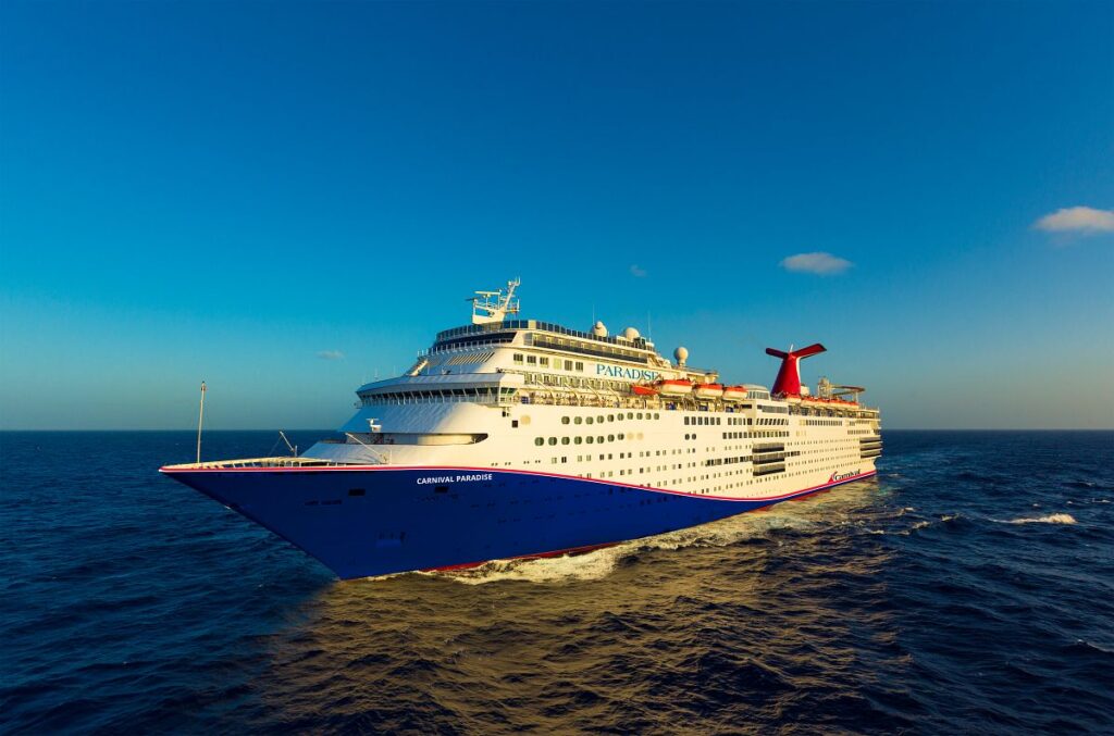 The Carnival Paradise cruise ship, bathed in the golden light of the setting sun, cruises the open sea, its blue hull and red funnel vibrant against the expansive sky and sea.