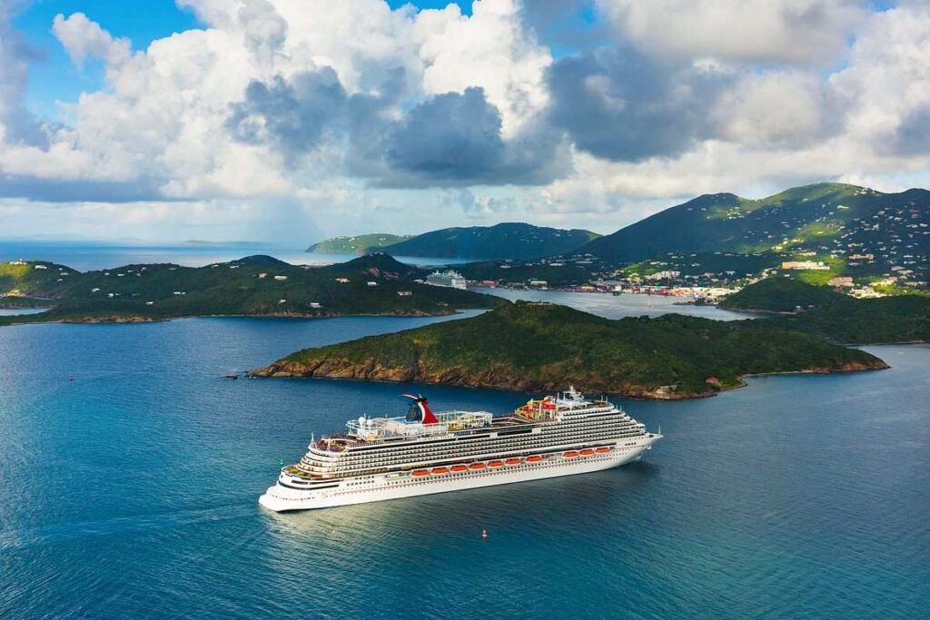 Carnival Vista sails near lush green islands under a sky dotted with clouds. The surrounding water is calm, reflecting the tropical scenery and the leisurely voyage of the cruise liner.
