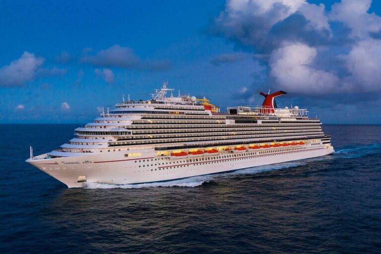 The Carnival Vista cruise ship sails on a serene blue ocean, its multiple decks and distinctive red funnel standing out against a backdrop of scattered clouds in a bright sky.