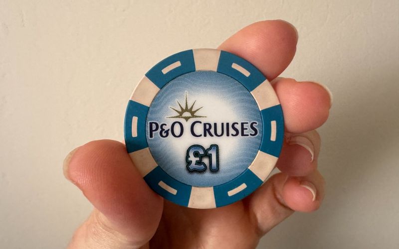 A hand holding a blue and white £1 P&O Cruises casino chip against a plain light-colored background.