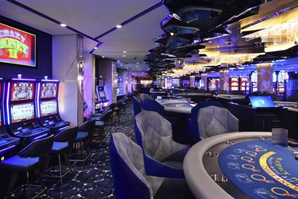 This image showcases the casino aboard a Celebrity cruise ship, featuring a vibrant and luxurious gaming environment. The casino is equipped with slot machines illuminated by colorful LED lights, sleek gaming tables with plush chairs, and a reflective ceiling that enhances the modern aesthetic. The ambiance combines excitement and sophistication, offering guests a thrilling onboard entertainment experience.