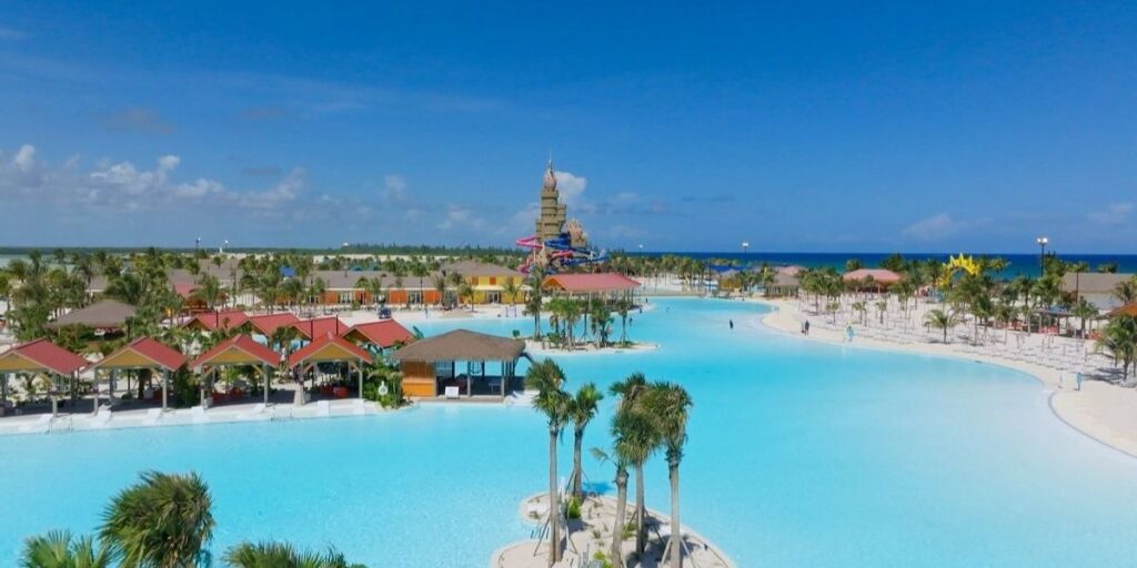 Aerial view of Celebration Key, Carnival's private destination, showcasing a sprawling turquoise lagoon surrounded by palm trees, colorful cabanas, and sandy walkways. In the distance, a large sandcastle-themed water slide structure rises near the shoreline, with the ocean visible on the horizon.