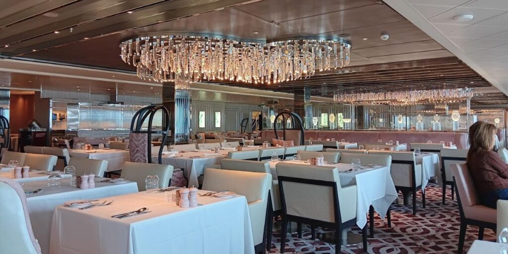 An elegant dining room on the Celebrity Apex cruise ship, featuring white tablecloths, neatly arranged table settings, and plush seating. The room is adorned with a large, sparkling chandelier, creating a luxurious atmosphere for guests.