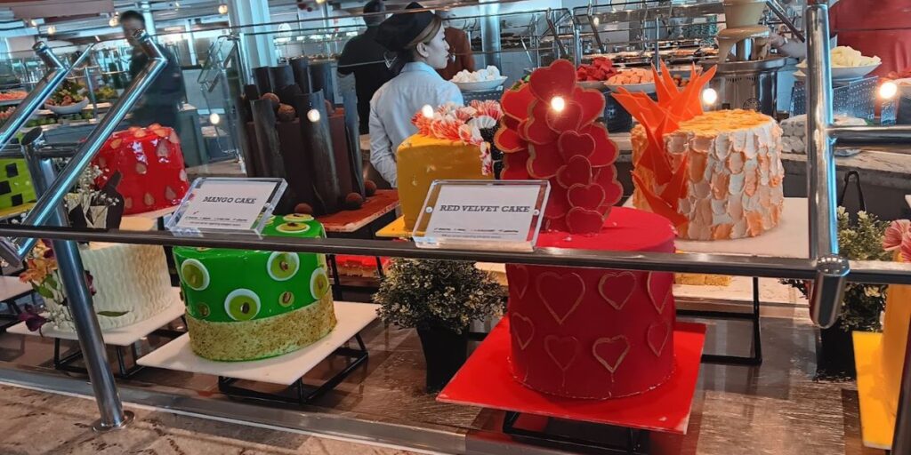 A colorful display of various cakes in the buffet area on the Celebrity Apex cruise ship, including a mango cake with green and yellow decorations and a red velvet cake adorned with heart-shaped designs. The cakes are set behind a glass barrier, highlighting the ship's diverse and artistic dessert offerings.