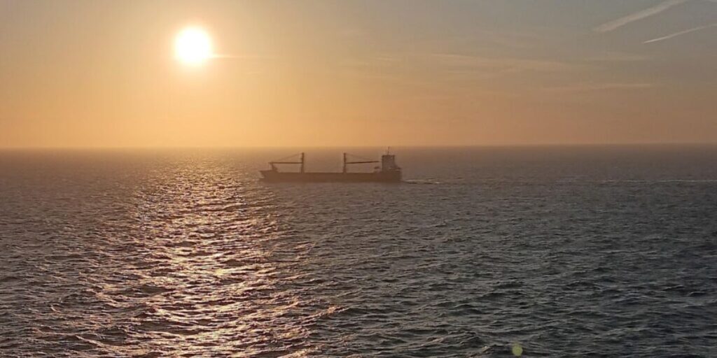A serene ocean sunset with a large cargo ship sailing in the distance, captured from the Celebrity Apex cruise ship. The sun casts a golden glow over the water, creating a peaceful and picturesque scene.