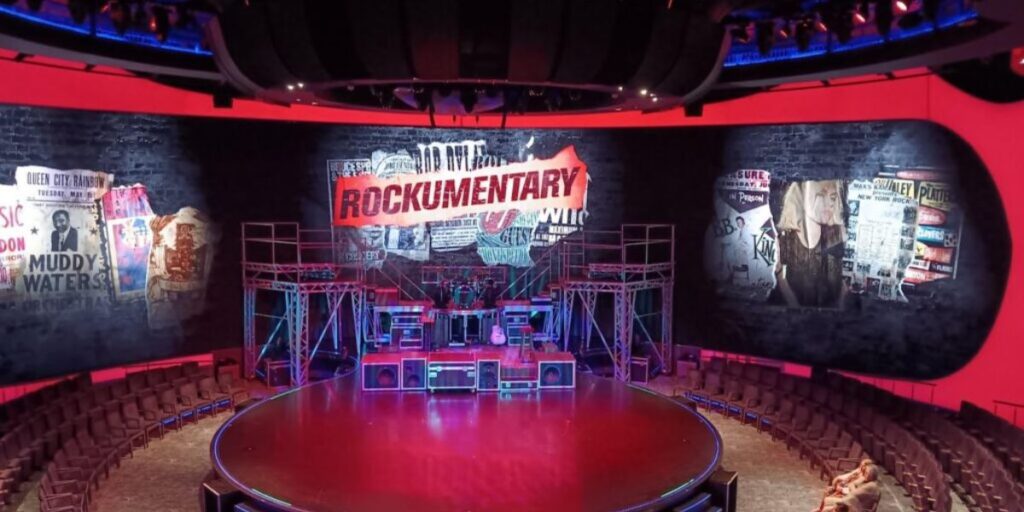 The theater on the Celebrity Apex cruise ship, set up for a show titled "Rockumentary." The stage is surrounded by a large circular seating area, and the backdrop features various rock music posters and memorabilia. The atmosphere is set for an exciting and immersive rock-themed performance.